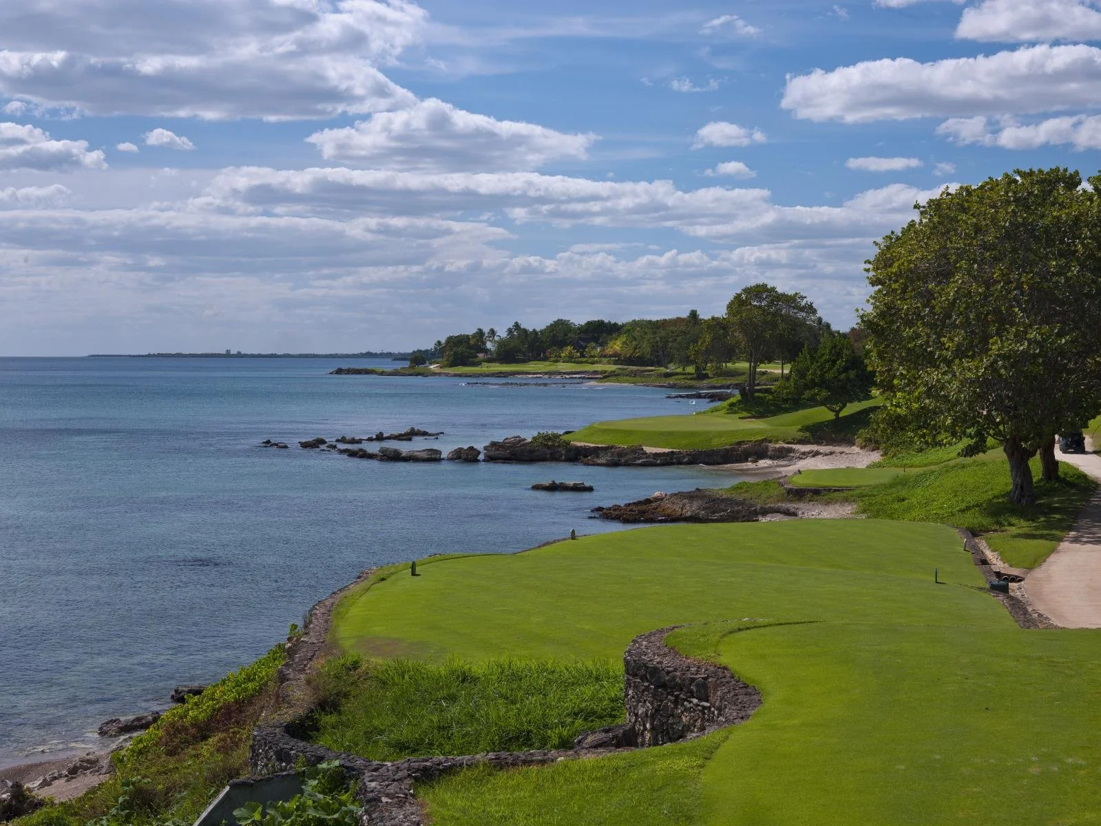 A scenic view of a golf course by the ocean with green grass, rocks along the shoreline, trees, and a partly cloudy sky.