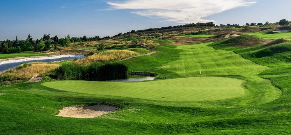 A scenic golf course with well-maintained green grass, sand bunkers, water hazards, and rolling hills in the background.
