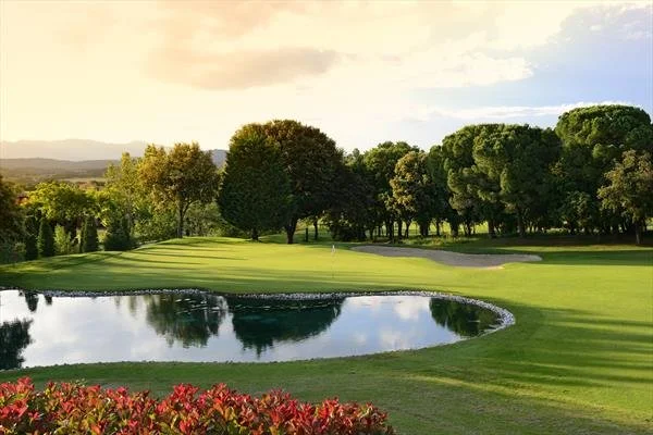 A golf course with a water hazard, lush green fairways, and trees in the background under a partly cloudy sky at sunset.