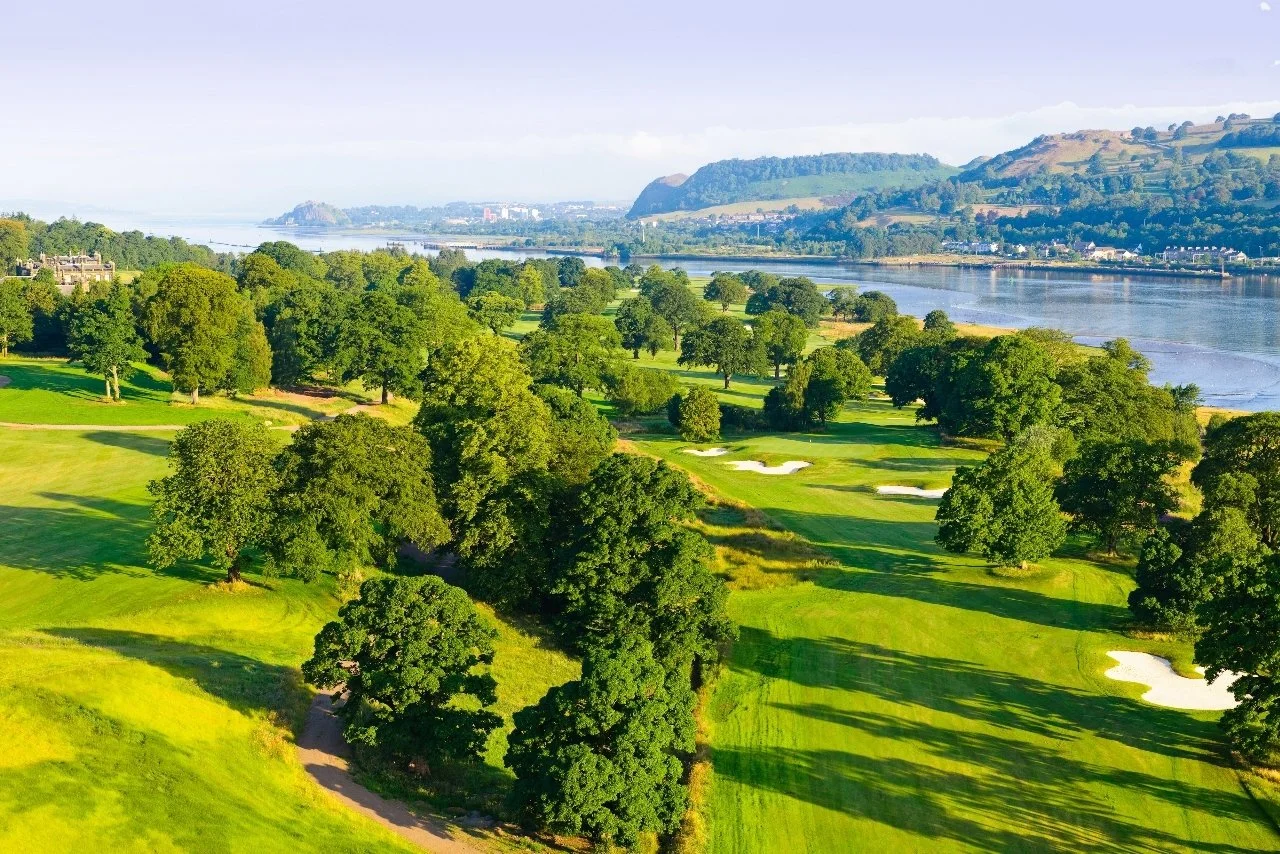 A scenic view of a golf course with lush green trees, water bodies, and rolling hills in the background.