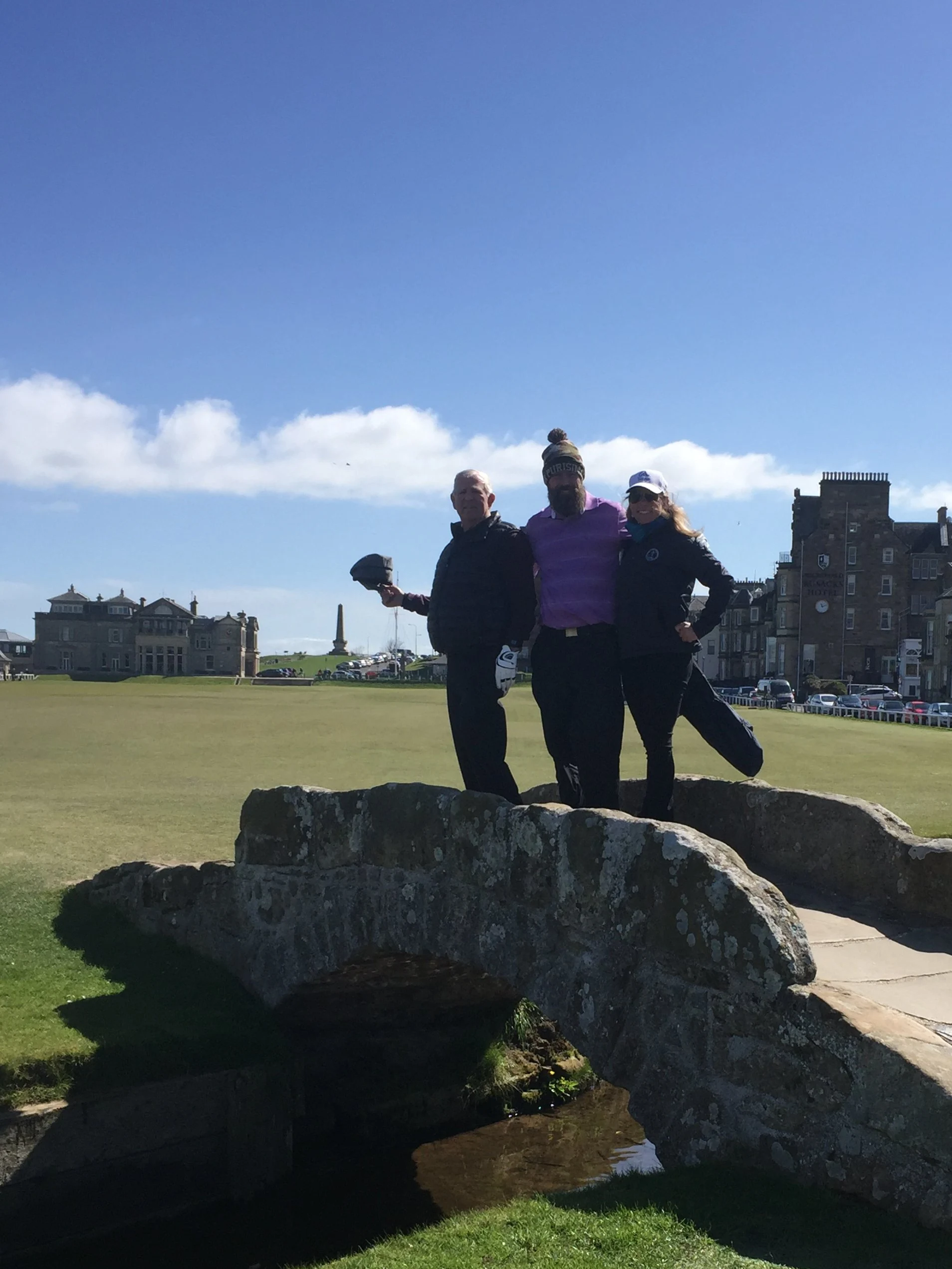 Three Clients standing on the Swilcan bridge at the old course St Andrews under a blue sky with scattered clouds.