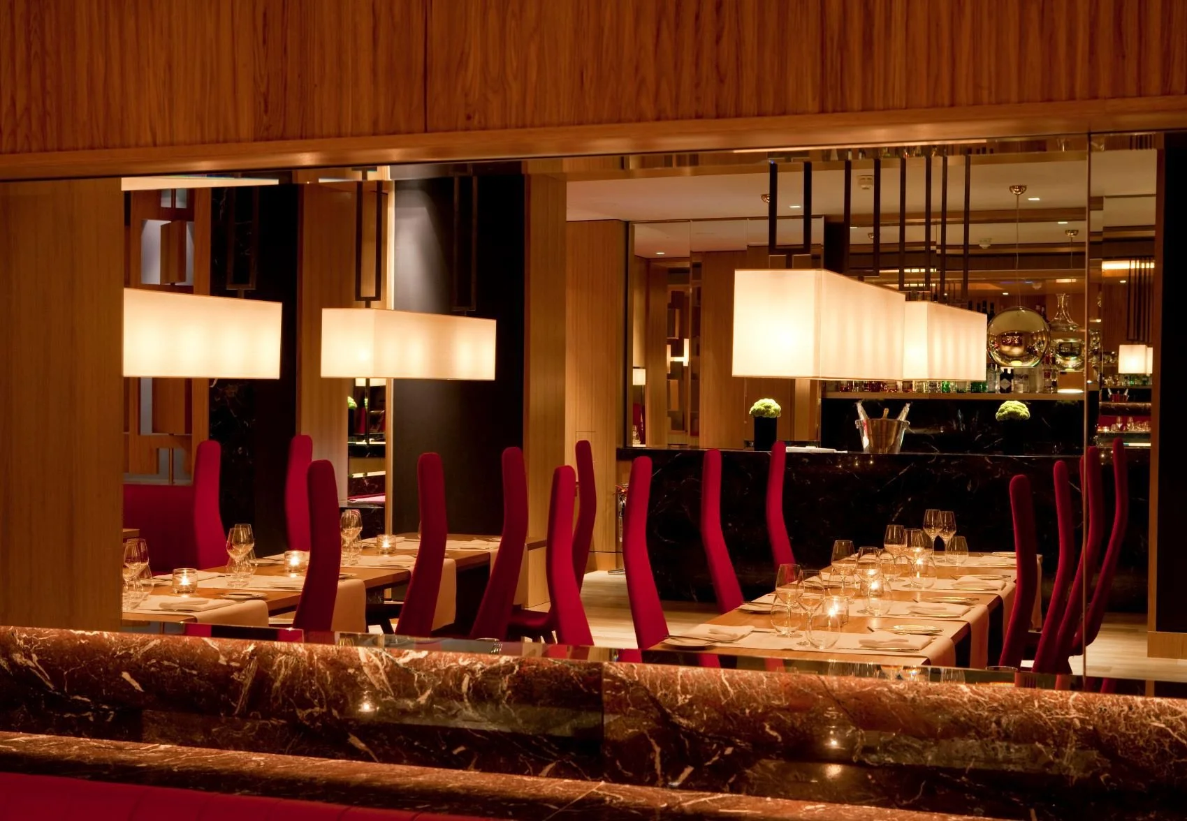 Interior of a upscale restaurant with wooden and marble decor, red high-back chairs, and dim lighting from large rectangular pendant lights.