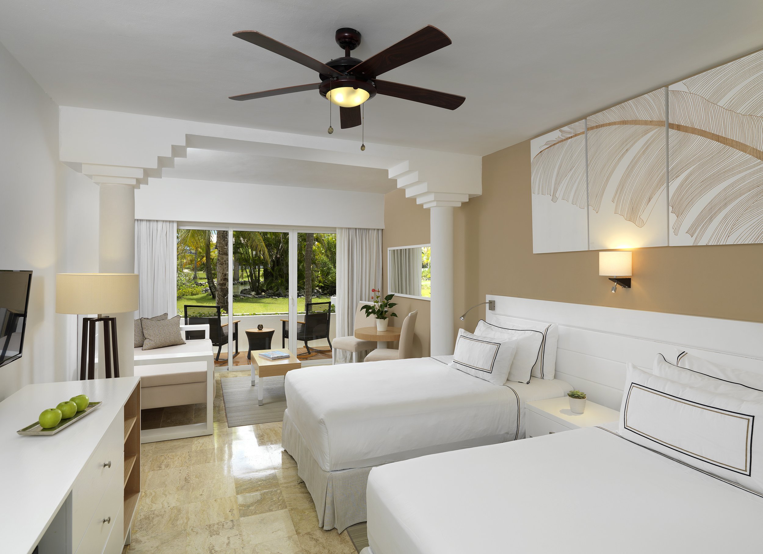 A bright hotel room with two white beds, a small desk with a mirror, a seating area with a white armchair and a sofa, a ceiling fan, and a sliding glass door leading to a patio with green trees and grass.