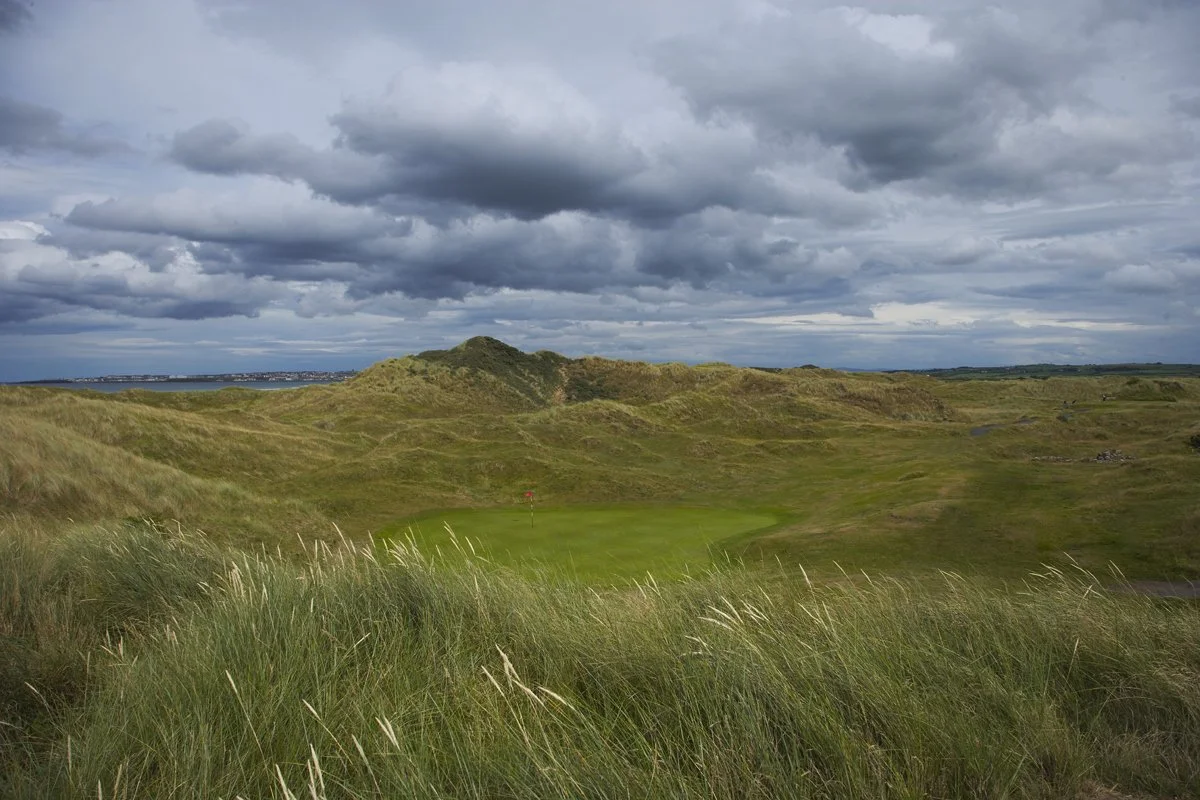A scenic golf course with lush green grass and a putting green in the middle, surrounded by rolling grassy hills and a cloudy sky.