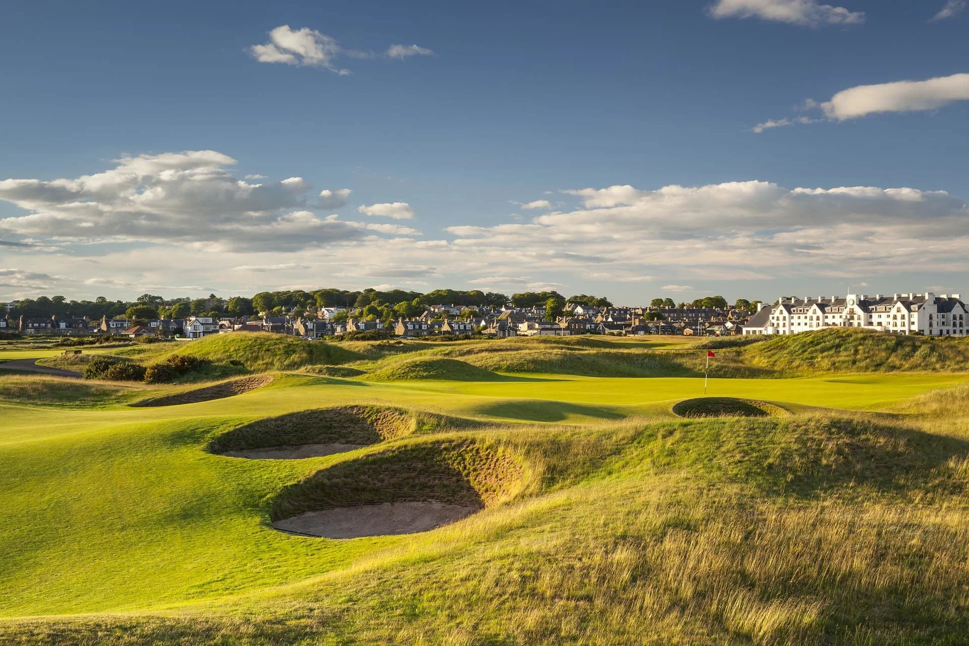 A scenic view of a golf course with sand bunkers, a green with a flag, and a distant town with white residential buildings under a partly cloudy sky.