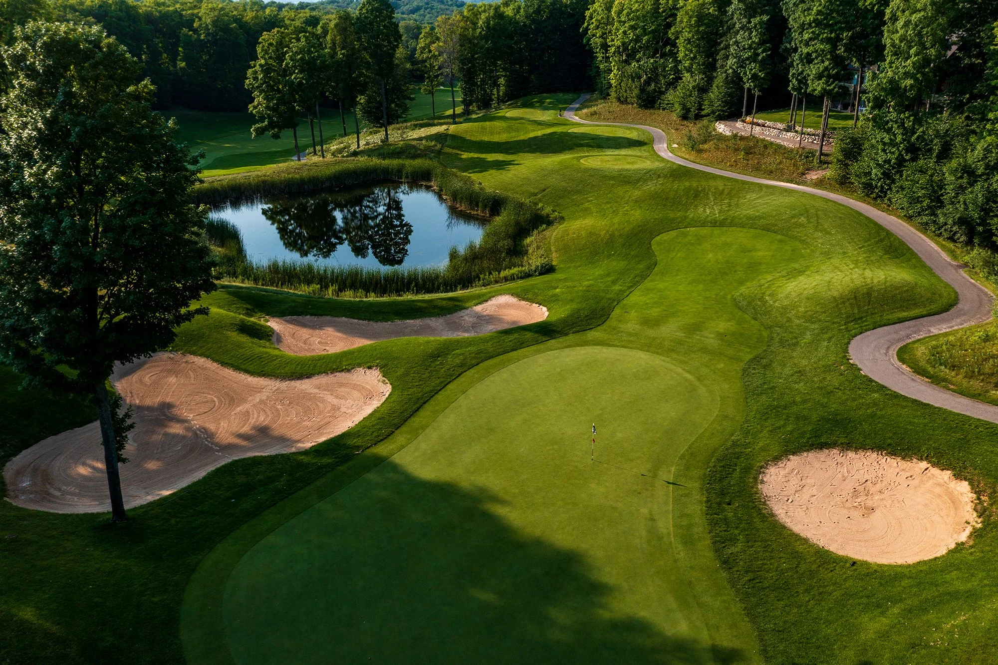 A golf course with a putting green, sand bunkers, a water hazard, and lush green trees in the background.