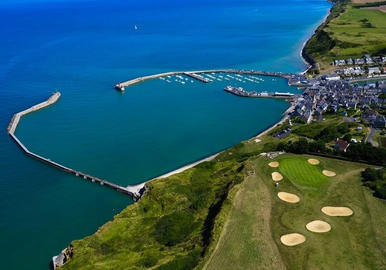 Aerial view of a harbor with a curved pier, a small coastal town, and a golf course with multiple sand bunkers near the shoreline.