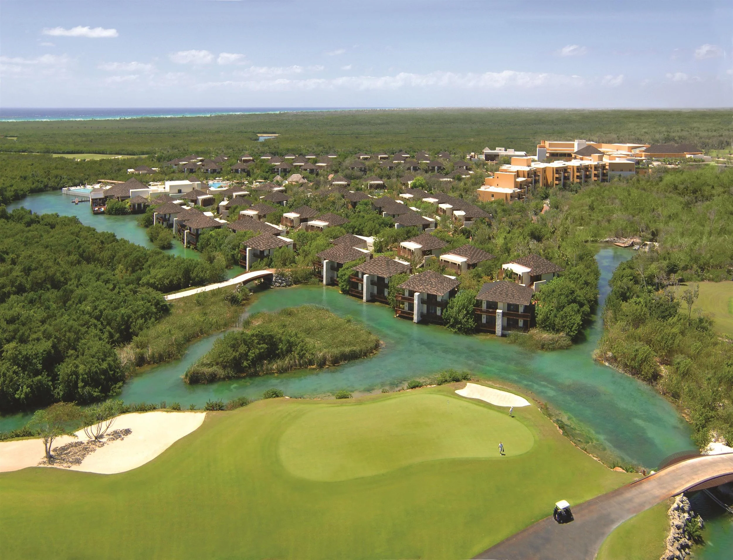 Aerial view of a luxury resort with multiple modern villas, surrounded by lush greenery, a winding river, and a golf course, with the ocean in the background.