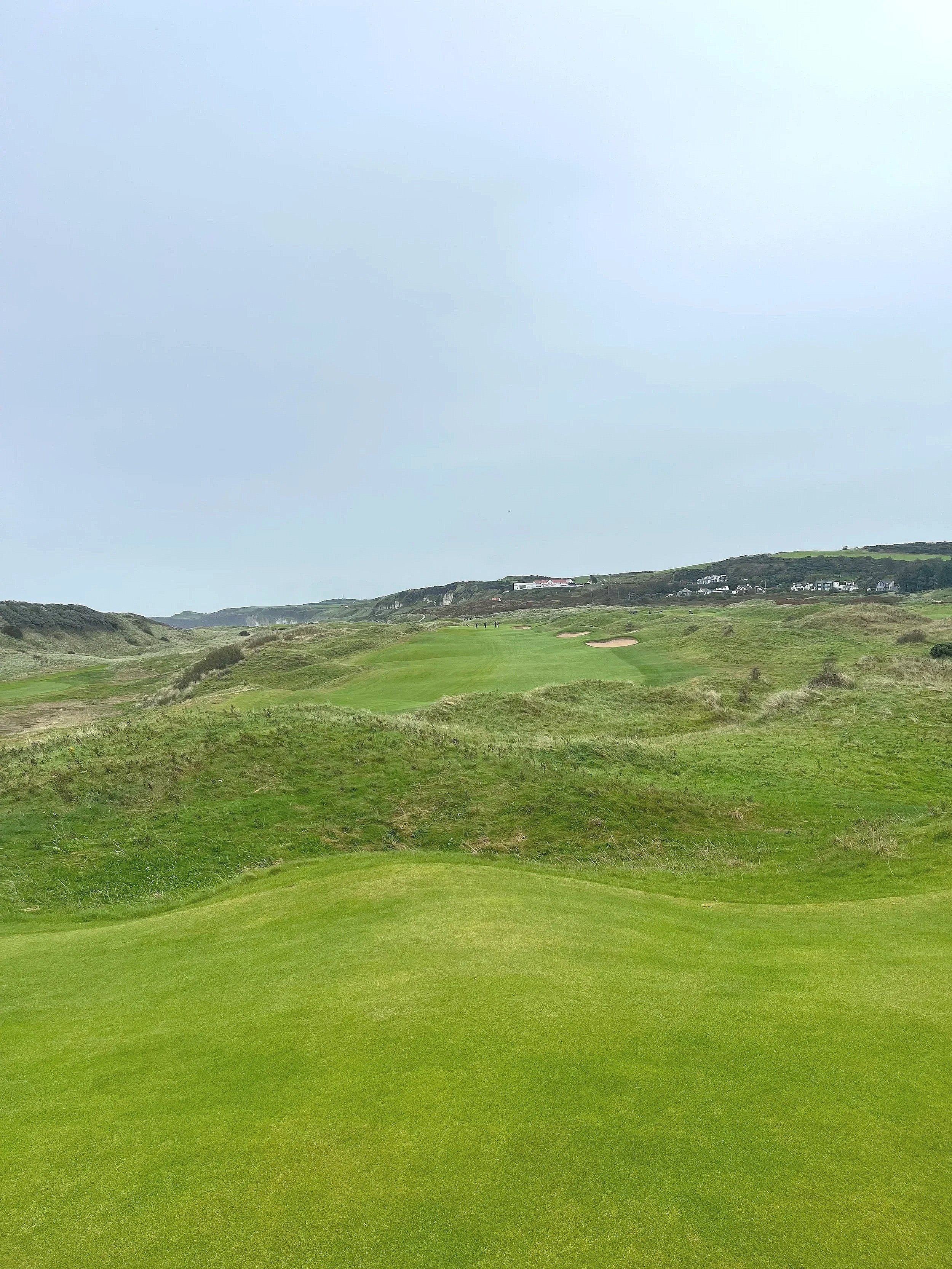 A golf course with lush green fairways, sand bunkers, and hilly terrain under a cloudy sky.