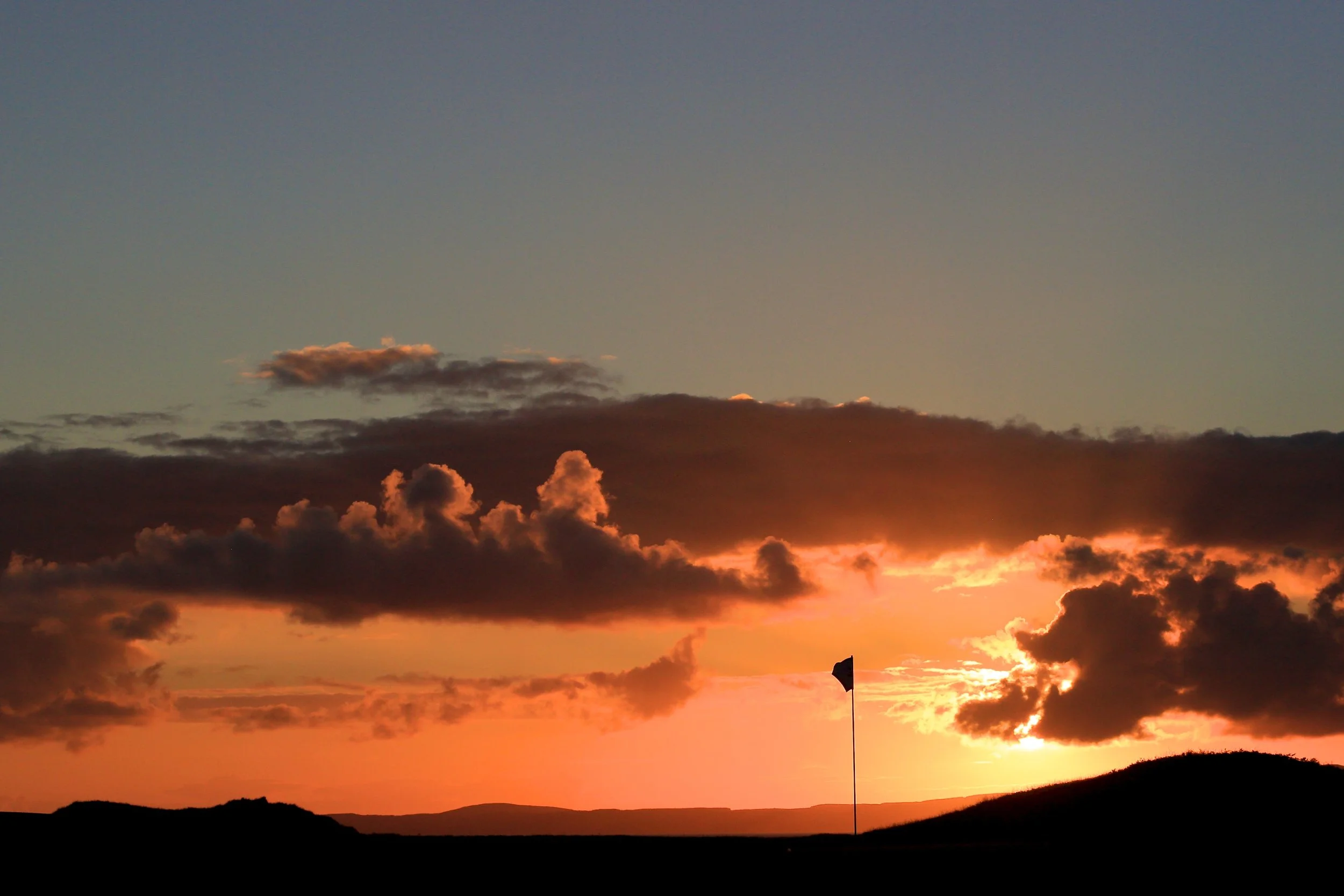 Sunset over a golf course with a flagstick and golf hole, silhouetted hills, and dramatic clouds in the sky.