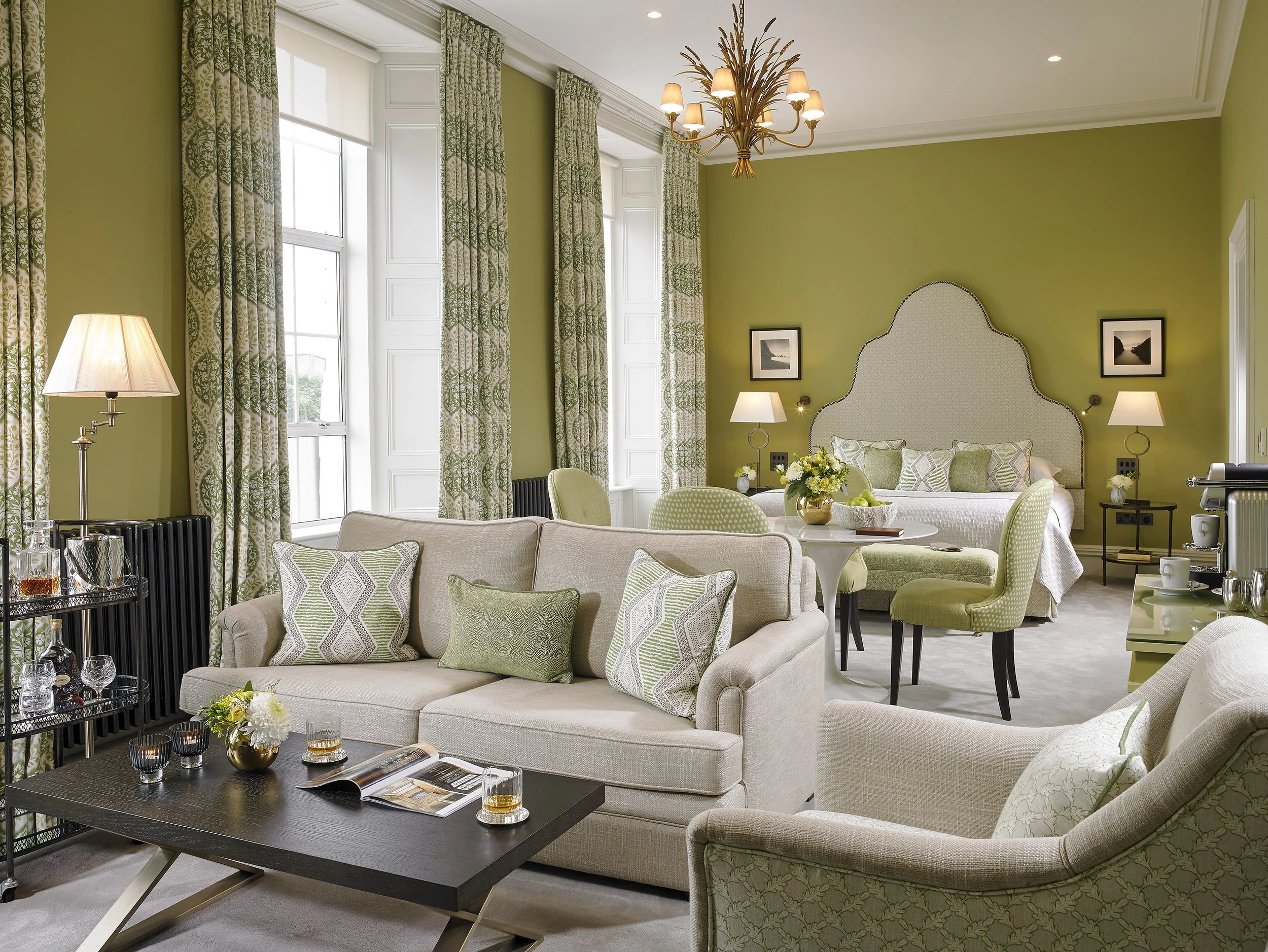 A spacious living room with green walls, a beige sofa with decorative pillows, a black coffee table with glasses and flowers, green and cream chairs, a round dining table, and a bed in the background with matching green and white decor.
