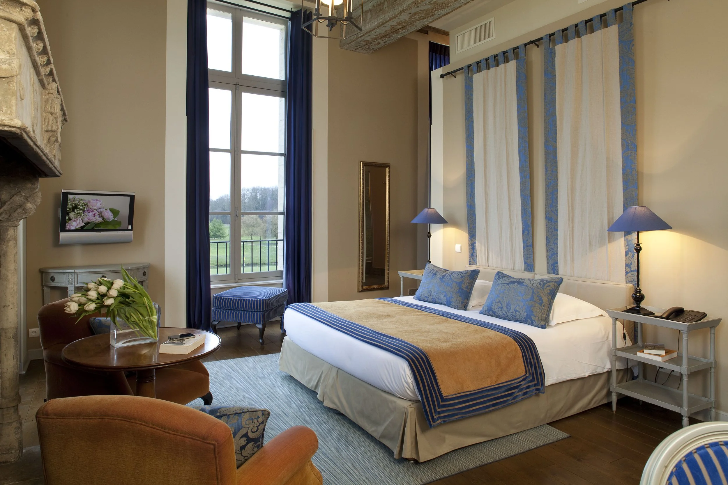 A hotel room with a large bed, blue pillows and a beige bedspread, lamps on nightstands, a window with blue curtains, a chair, a small table with flowers, a mirror, and a small TV, with a view of a green landscape outside.