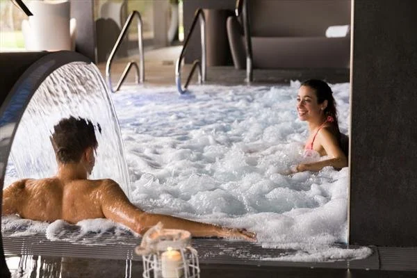 Two people relaxing in a hot tub with bubbling water, one woman smiling and sitting upright, and a man leaning on the edge with his back to the camera, on an indoor pool deck.