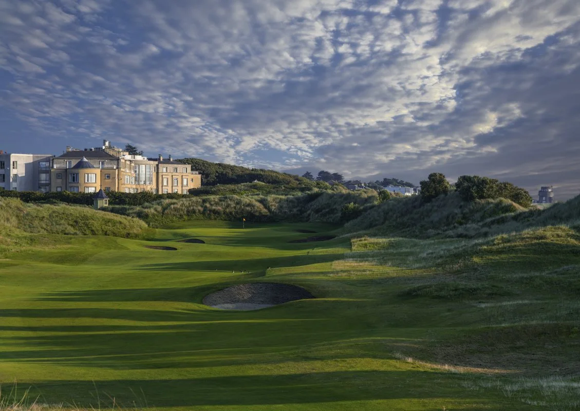 A scenic golf course with green fairways, sand bunkers, and a flag on the green, surrounded by rolling hills and residential buildings under a partly cloudy sky.
