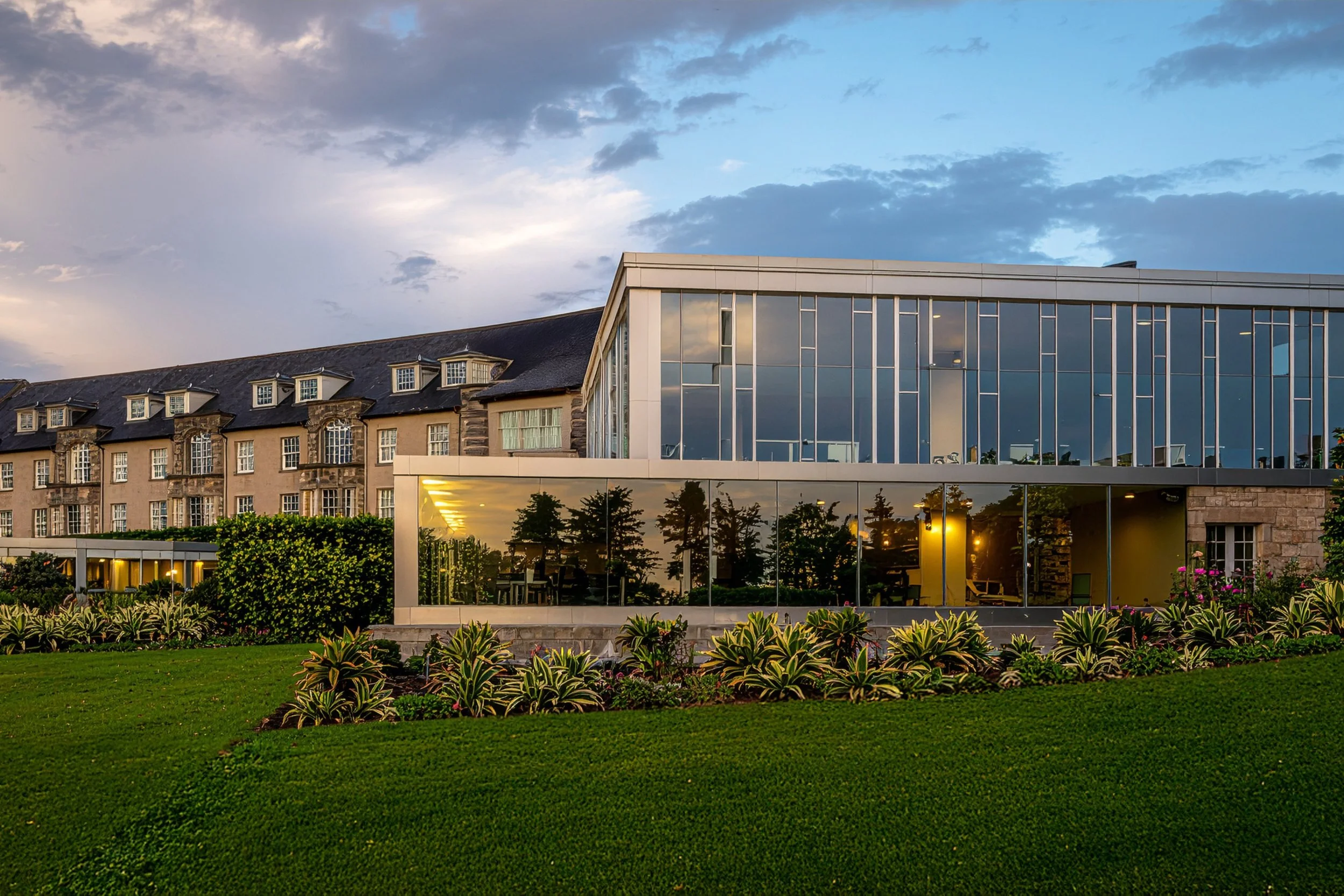 Modern glass building reflecting the sky and trees, with a landscaped green lawn and flower bed in the foreground, under a partly cloudy sky.