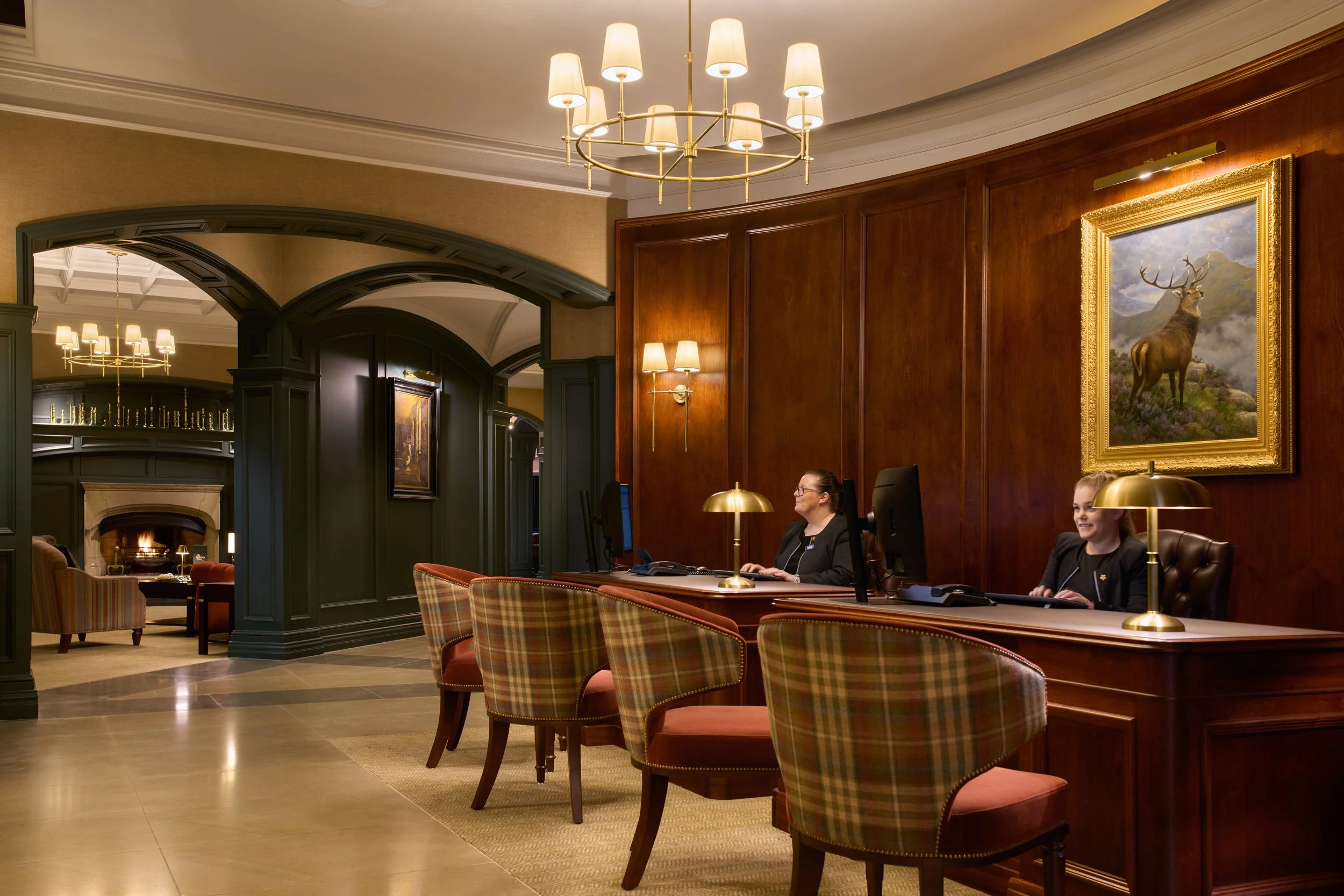A hotel lobby or reception area with two women working at reception desks with computers, wooden paneling, gold lamps, a framed painting of a deer, and chandeliers lighting the space.