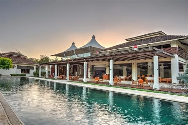 Luxury poolside area with lounge furniture and a building with a canopy roof, overlooking a swimming pool at sunset.