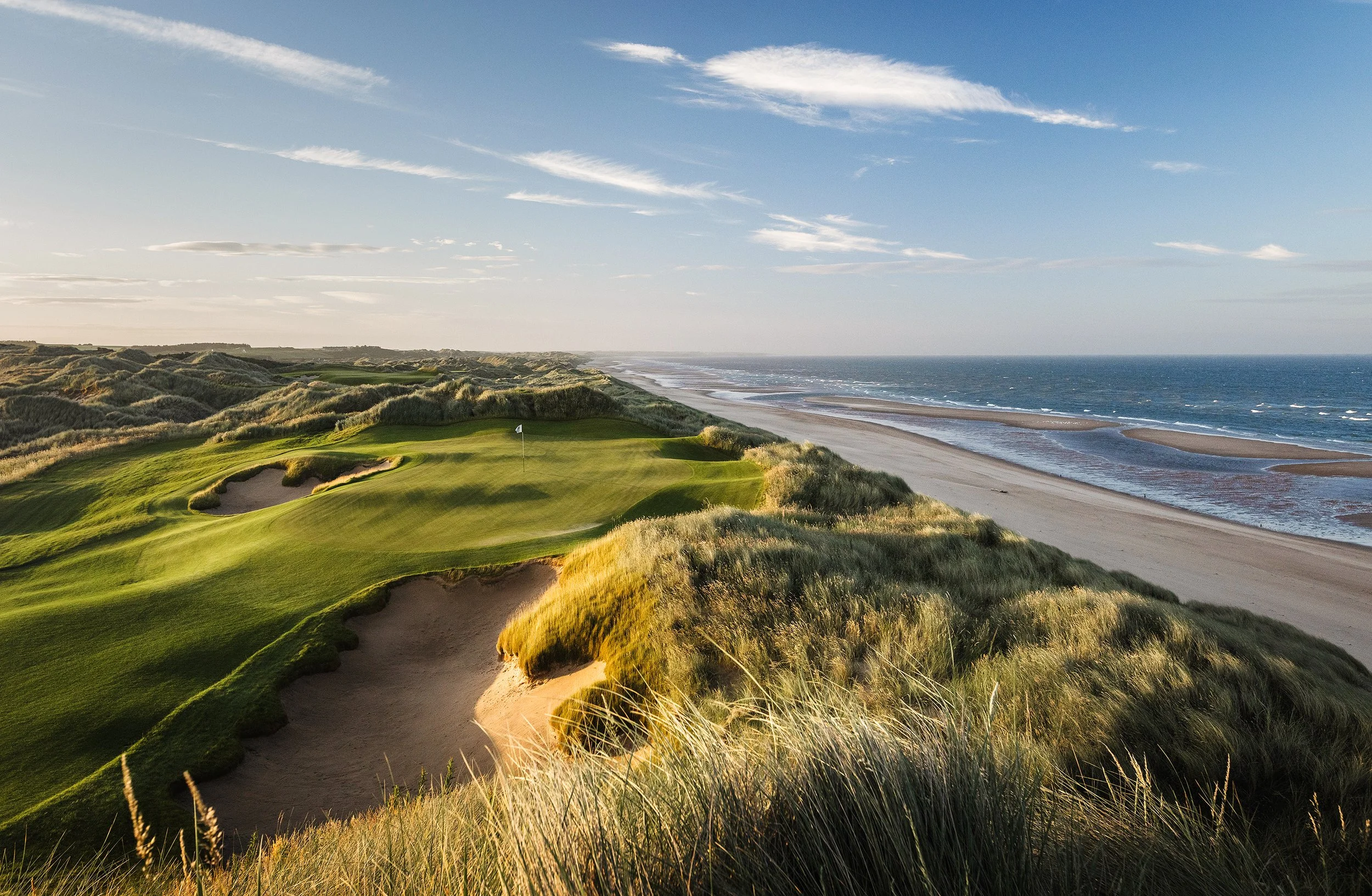 A scenic view of a golf course near the coastline, with green grass, sandy bunkers, and a flag on the green, overlooking the ocean under a clear sky.