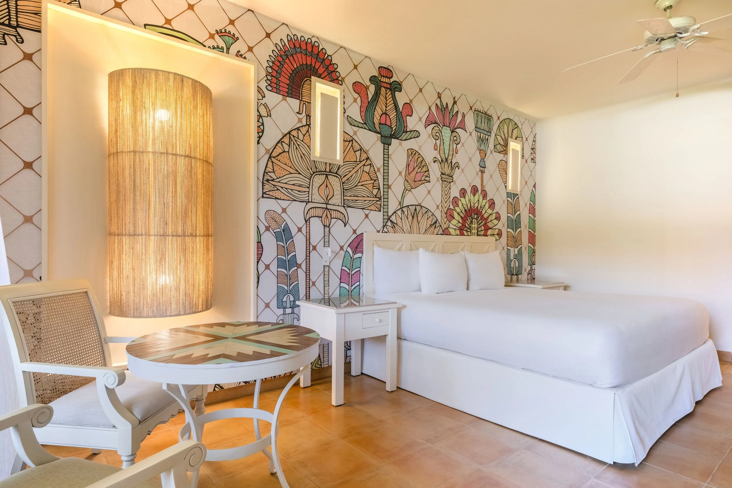 Hotel bedroom with white bed, colorful patterned wall art, and wooden furniture including a round table and chairs, illuminated by soft lighting.