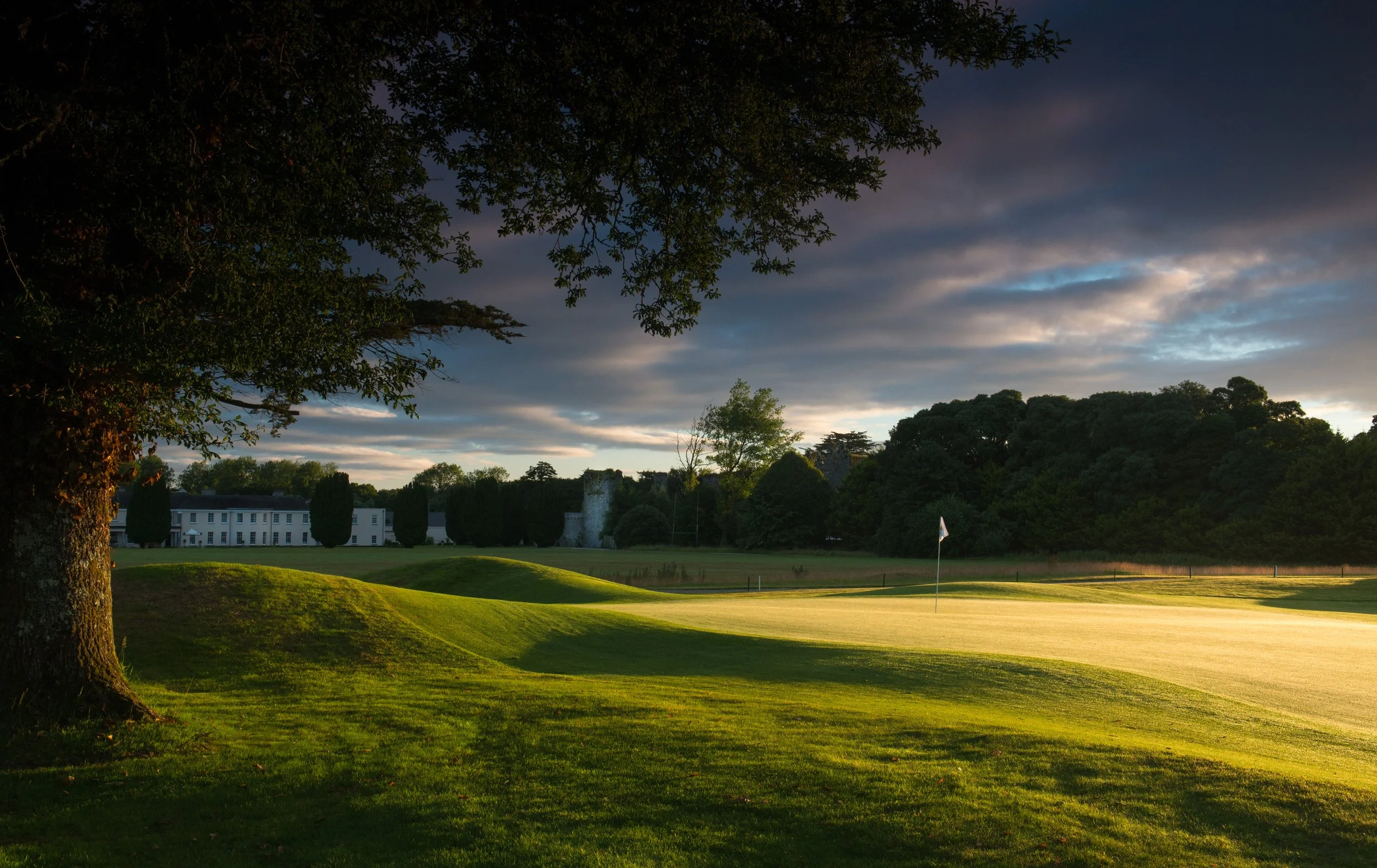 A golf course at sunset with a flag on the green, surrounded by trees and a large building in the background.