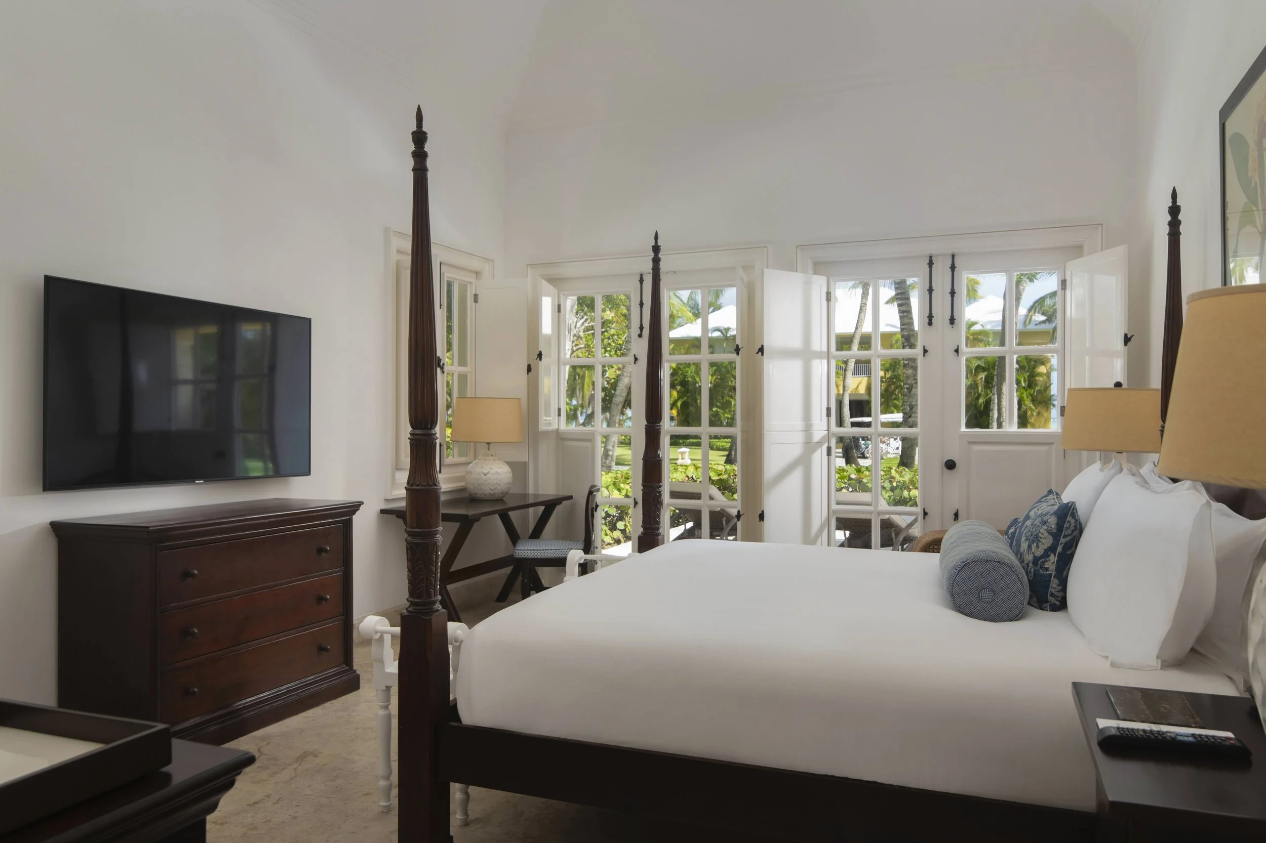 A bright, tropical hotel bedroom with a white bed, wooden four-poster frame, large windows and doors with a view of palm trees and lush greenery outside. There are nightstands with lamps, a dark wooden dresser, and a small desk with a chair. A flat-s