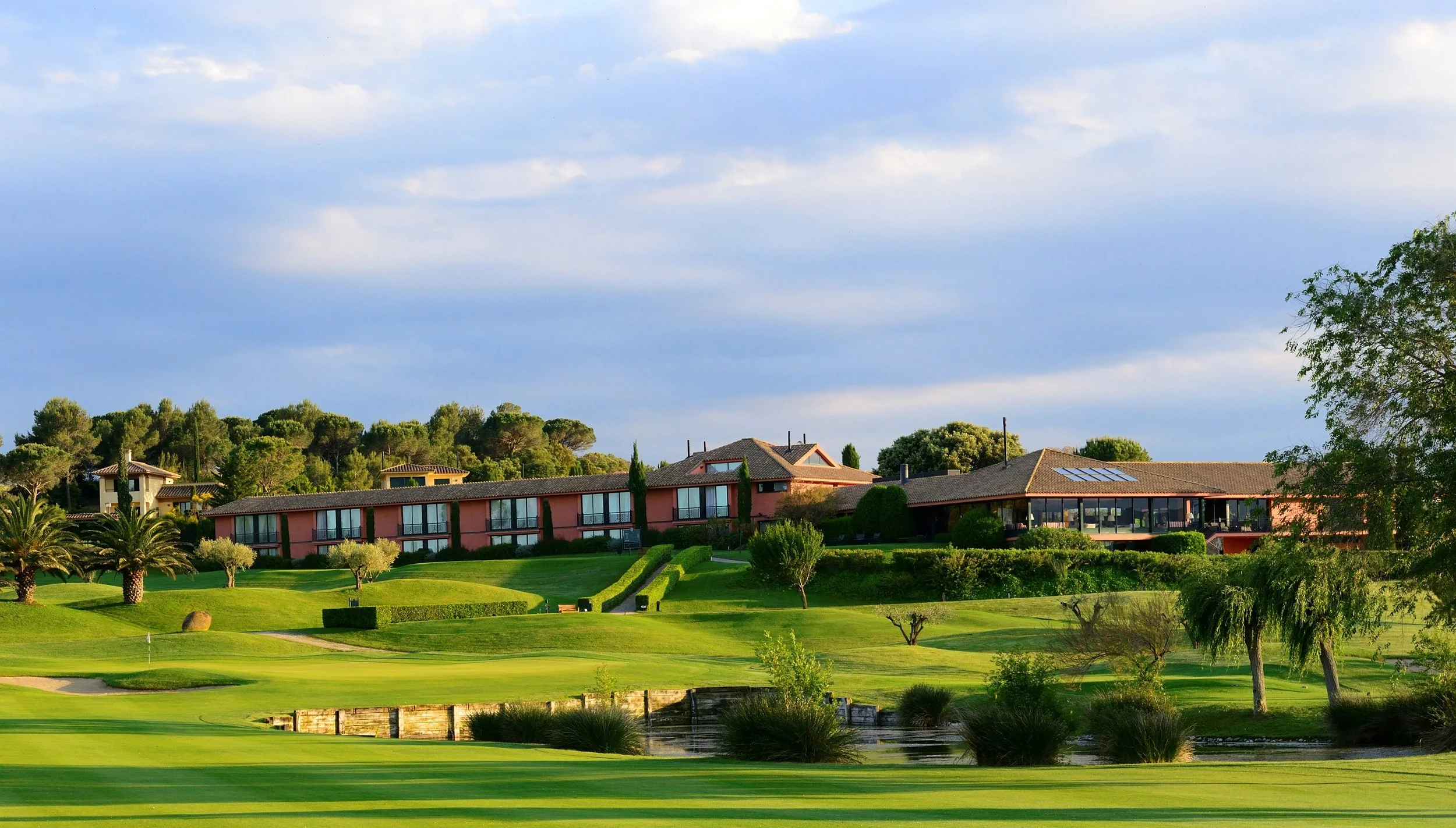 Panoramic view of a golf course with green fairways, trees, and a large building complex with terracotta roofs in the background, under a partly cloudy sky.