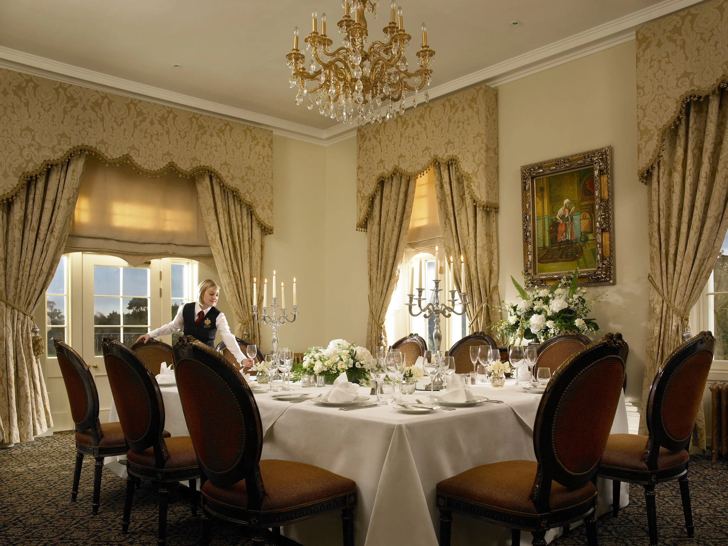 Elegant dining room set for a formal event with a large round table, white tablecloth, and floral centerpiece, surrounded by upholstered chairs. A woman in formal attire is arranging the table. The room features gold patterned curtains, a chandelier, candelabras, and framed artwork on the wall.