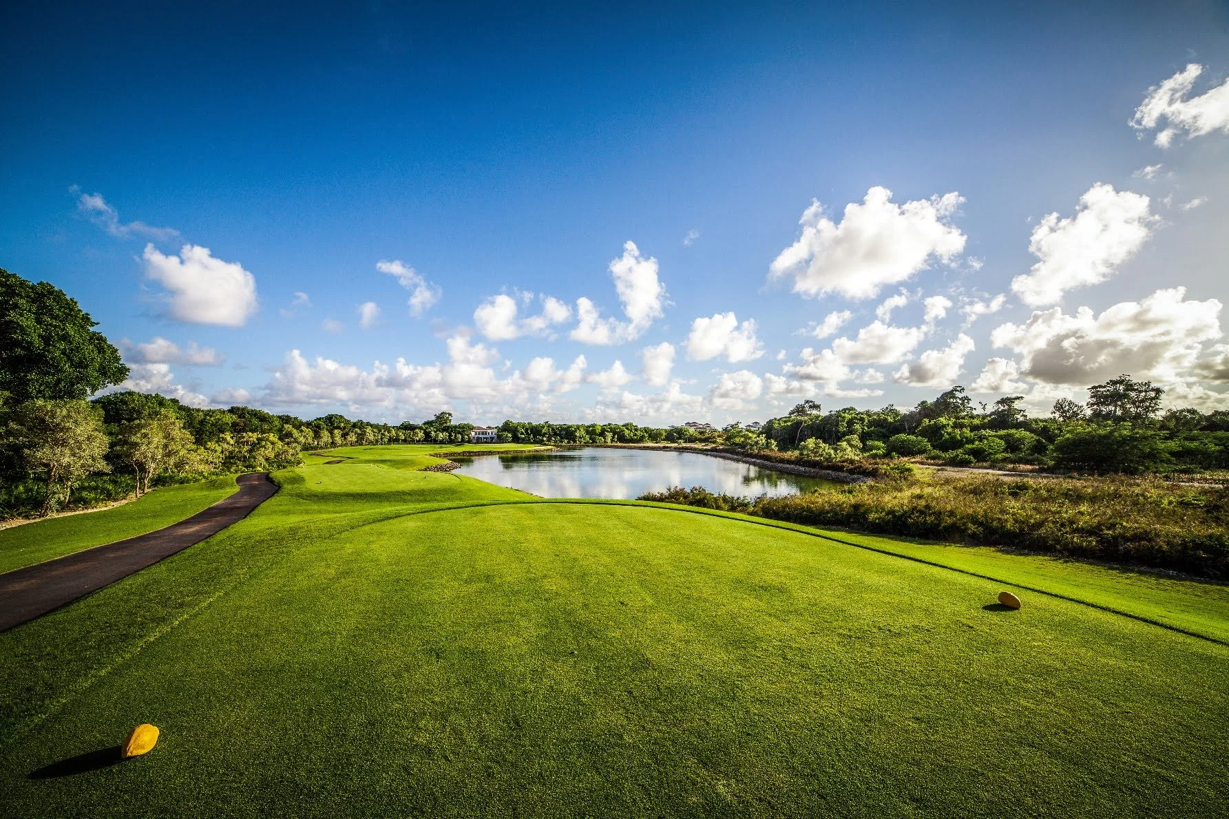 A golf course with green fairways, a water hazard, trees, and a blue sky with scattered clouds.