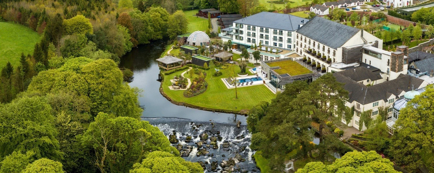 An aerial view of a large hotel or resort with a river running through a lush green landscape, featuring landscaped gardens, a swimming pool, and several small buildings amidst trees.