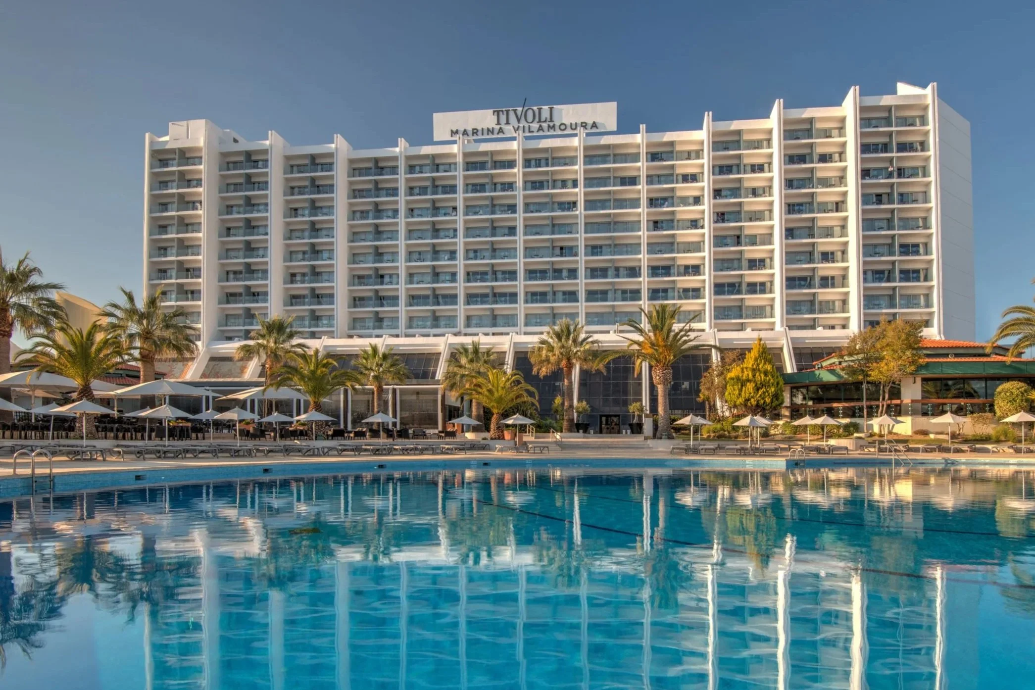 View of a large hotel building with the sign 'Tivoli Marina Vilamoura' on top, overlooking a swimming pool with lounge chairs and umbrellas, surrounded by palm trees.