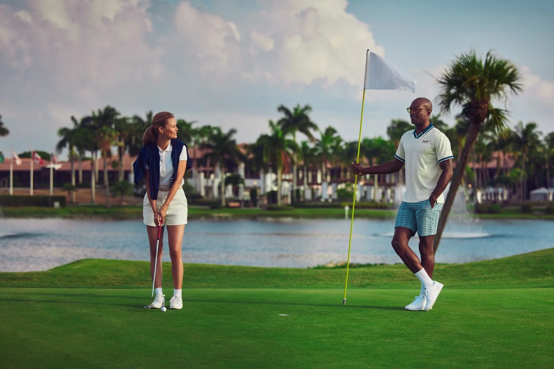 A man and woman preparing to play golf on a lush green course near a lake, with trees and buildings in the background.