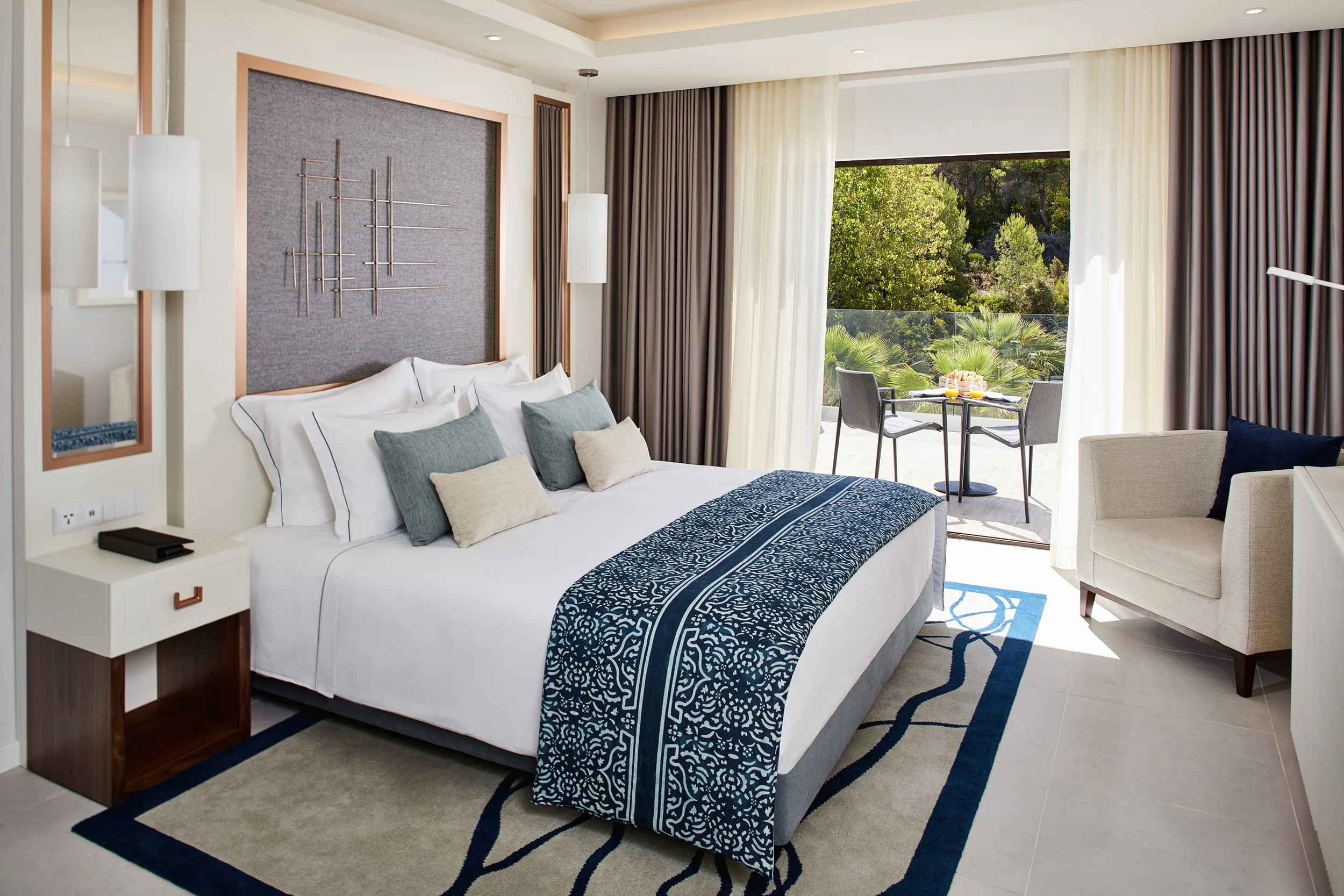 Hotel room with a large bed, decorative pillows, nightstand, armchair, and balcony with outdoor seating and greenery.