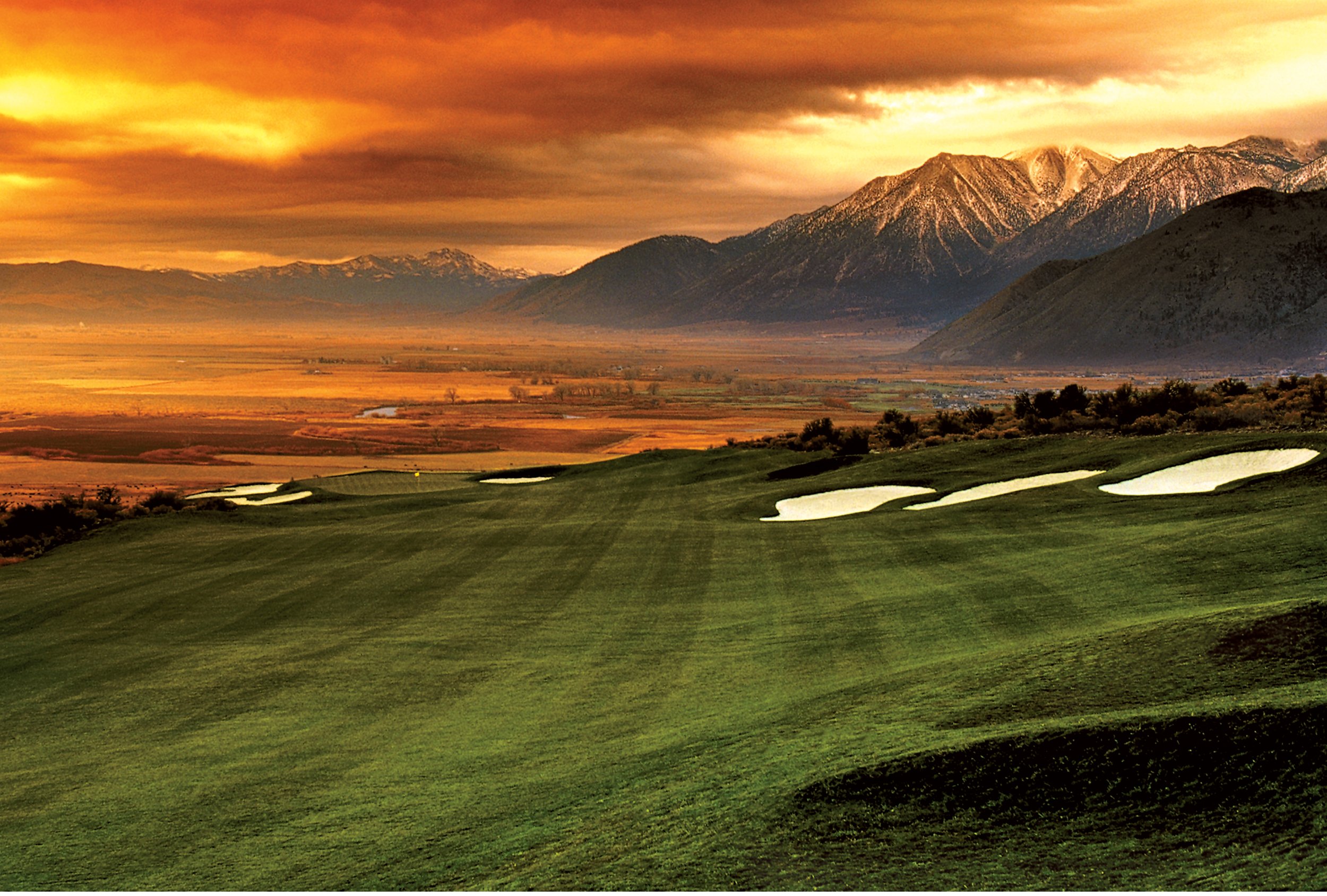 Golf course on rolling hills with sand traps, mountains in the background, and a colorful sunset sky.