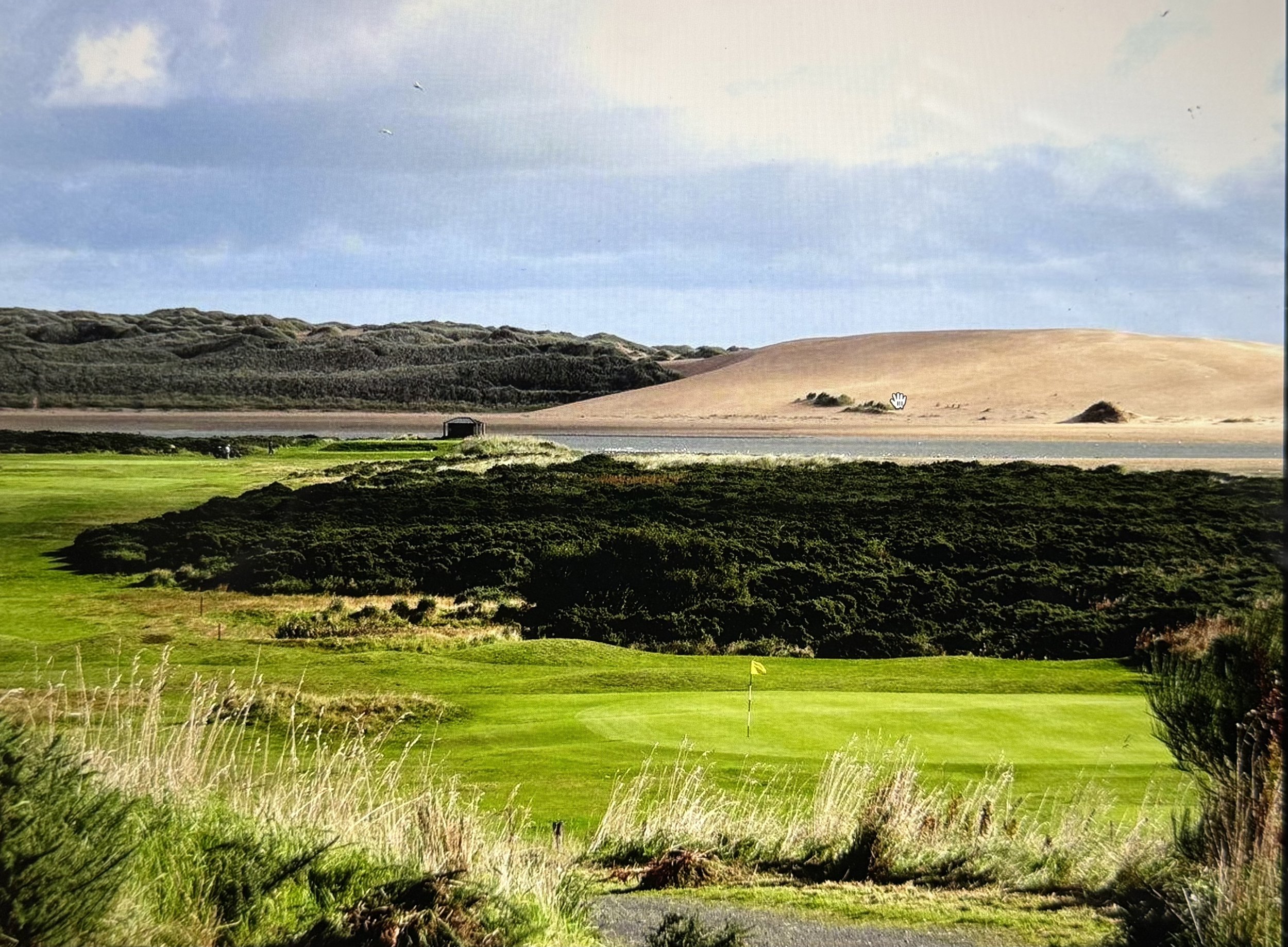 A scenic landscape with a golf course in the foreground, lush green grass, and a yellow golf flag. Behind the golf course, there are dark green bushes and trees. Further back, there is a body of water, with sandy dunes and a hill with sparse vegetation in the background under a partly cloudy sky.