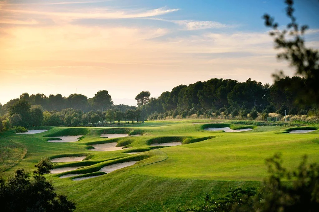 A scenic golf course at sunset with well-maintained green fairways, sand bunkers, and trees in the background under a partly cloudy sky.