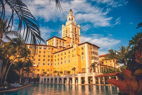 A large, yellow hotel with a clock tower, surrounded by palm trees, overlooks a pool under a blue sky.