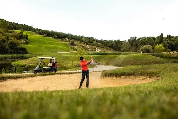 A person is playing golf in a sand bunker at a golf course, with a golf cart in the background and lush green hills surrounding the area.
