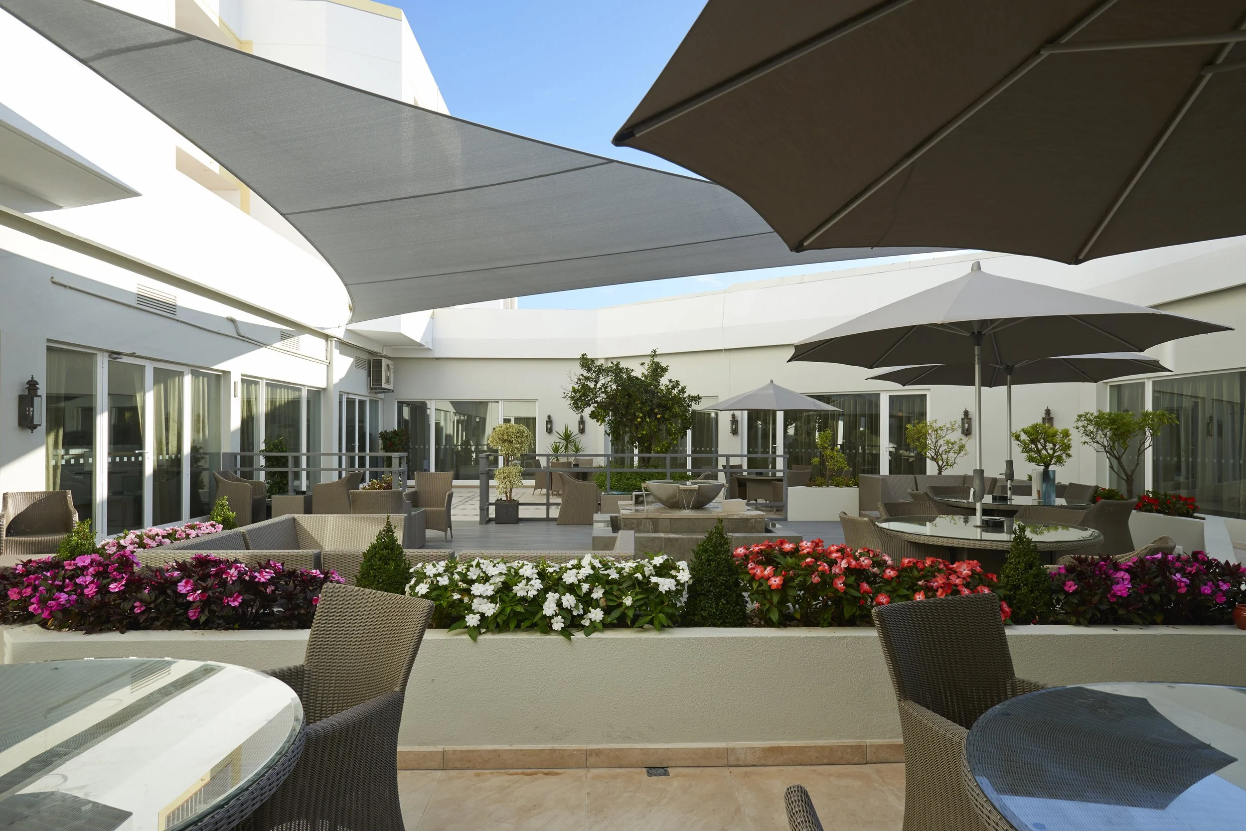 An outdoor terrace with several patio tables, chairs, and large umbrellas providing shade, surrounded by flower beds with pink, white, and red flowers, and green shrubbery, under a clear blue sky.