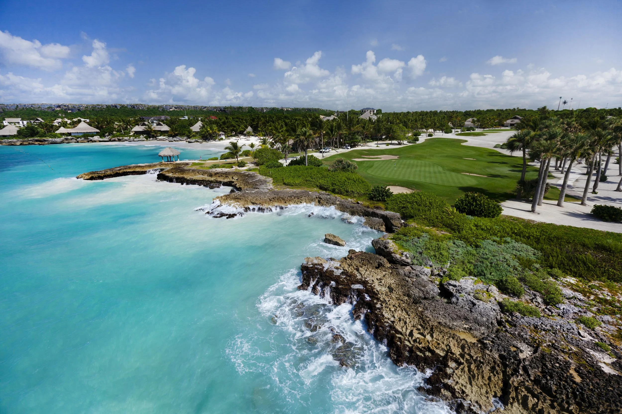 A coastal golf course with lush green grass, surrounded by palm trees, overlooking a bright turquoise ocean with rocky outcroppings and small thatched-roof structures on the beach.
