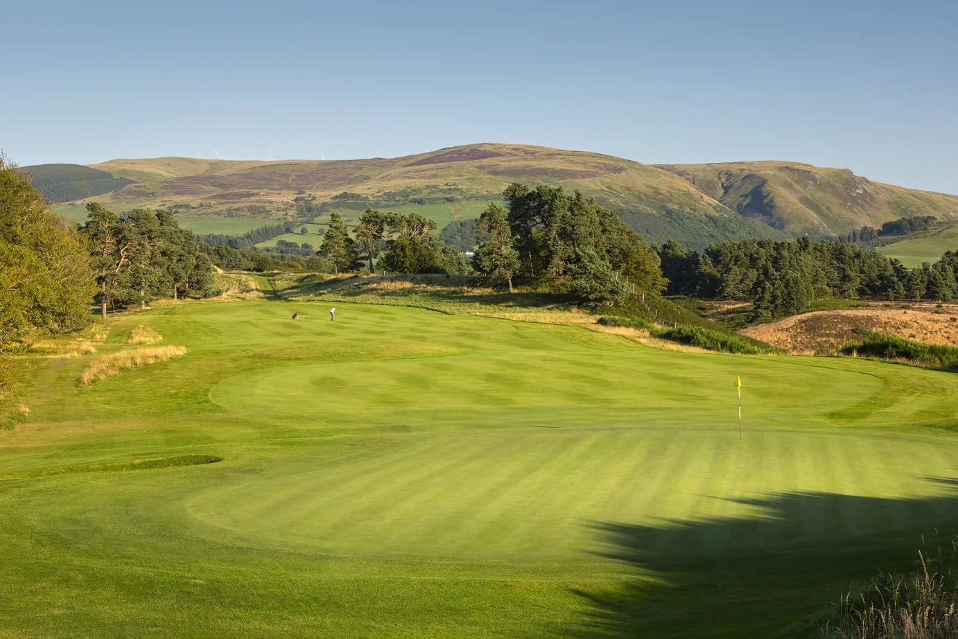 A lush green golf course with rolling hills and trees in the background, under a clear blue sky.