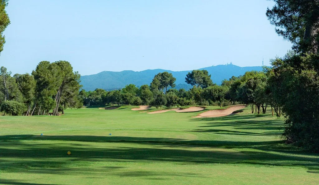 A golf course with green fairways, sand traps, and trees, with mountains in the background under a clear blue sky.