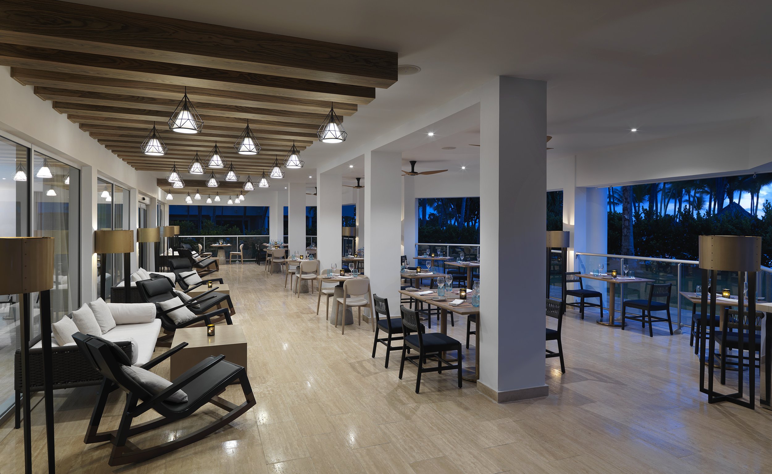 Interior of a modern restaurant or lounge with wooden floor, white walls, and large windows showing a tropical outdoor scene. The ceiling has wooden beams and hanging pendant lights. There are tables with chairs, and lounge seating with black cushion