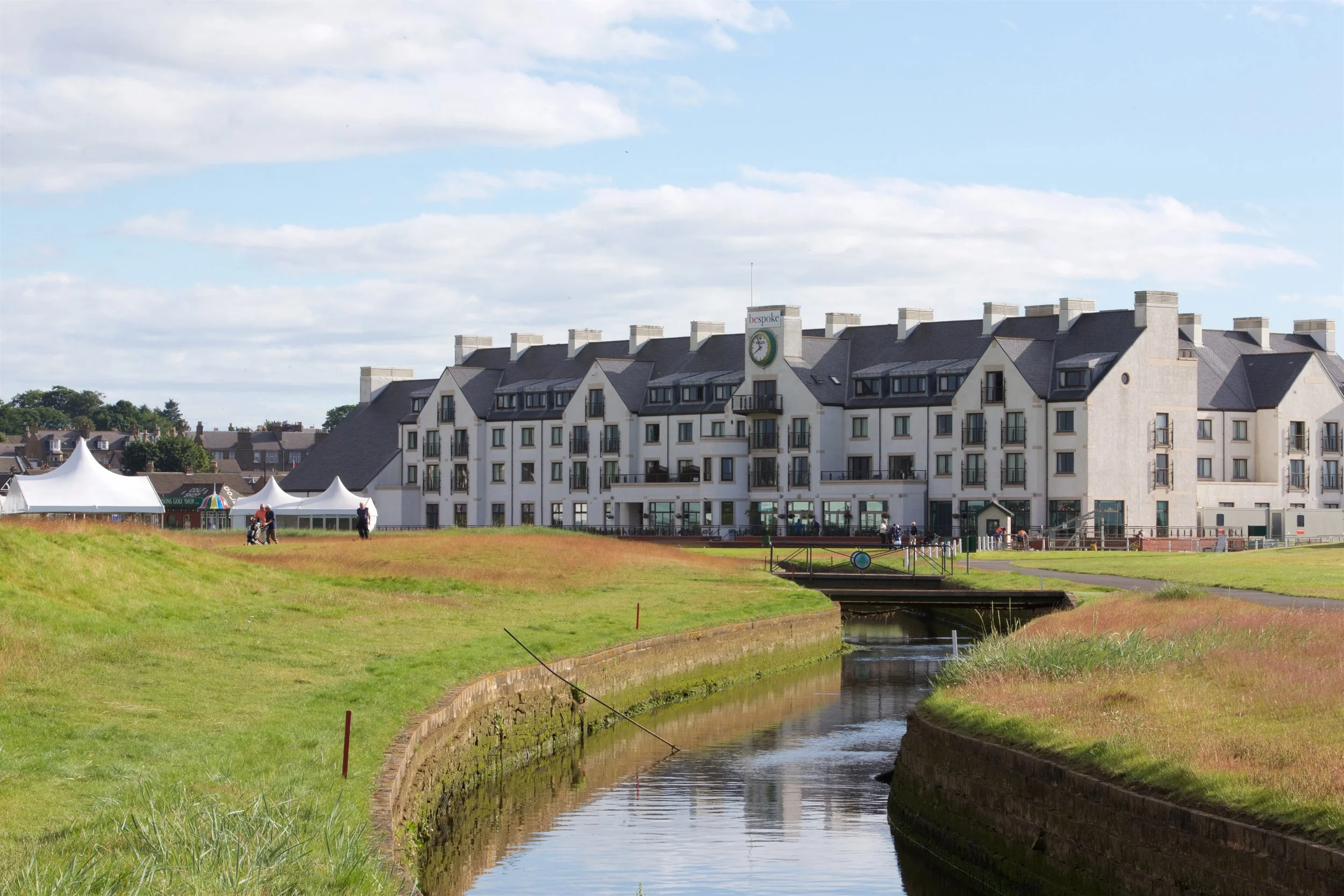 A large white modern hotel building near a small canal with grassy banks and a few people walking, under a partly cloudy sky.