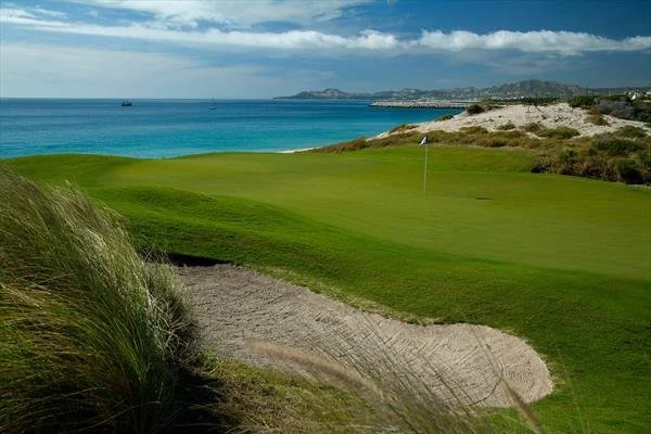 A scenic golf course near a beach with sand dunes, green grass, a flag, and ocean in the background under a partly cloudy sky.