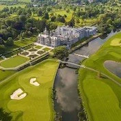 Aerial view of a large, historic castle-like building surrounded by a golf course with sand traps, water hazards, and lush green fairways, with trees and fields extending into the distance.