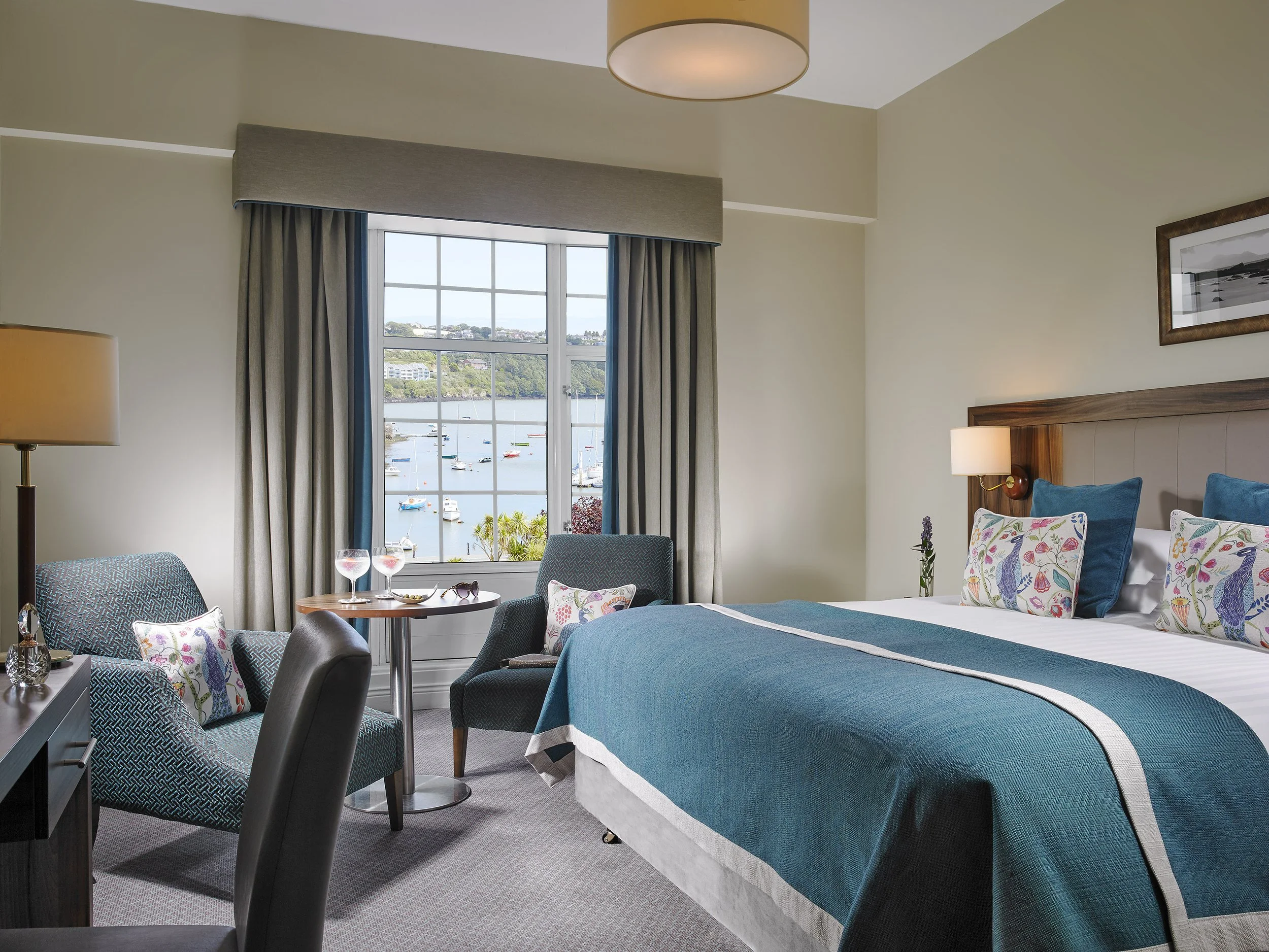 A hotel room with a large window overlooking a marina with boats, a bed with blue and white bedding, two chairs with decorative pillows, and a small table with wine glasses.