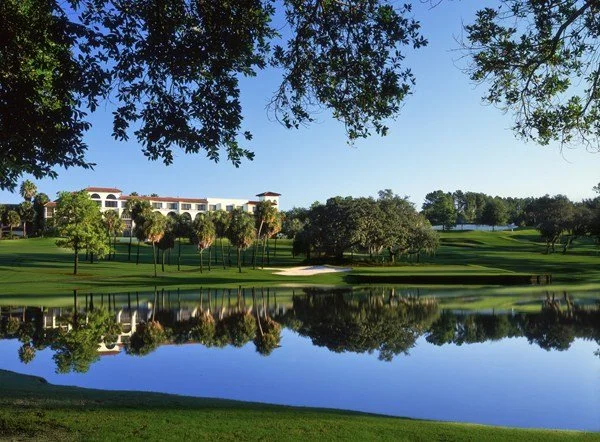 A lakeside golf course with trees and a large white resort building in the background under a clear blue sky.