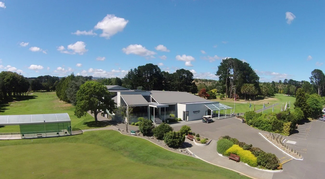 An aerial view of a building with a golf course, trees, and a sunny blue sky with scattered clouds.
