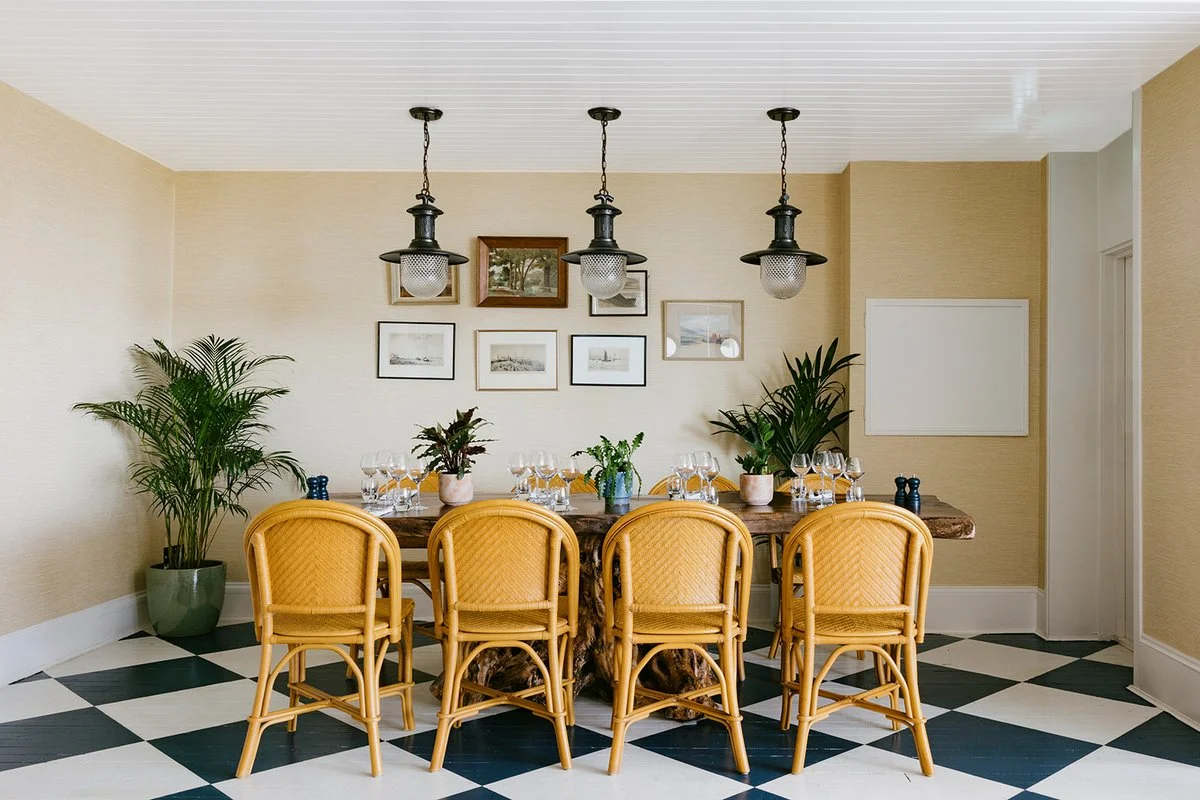 Dining room with six yellow wicker chairs around a rustic wooden table set with wine glasses and potted plants. Decorative framed pictures on the wall and large potted plants in the corners. Black and white checkered floor.