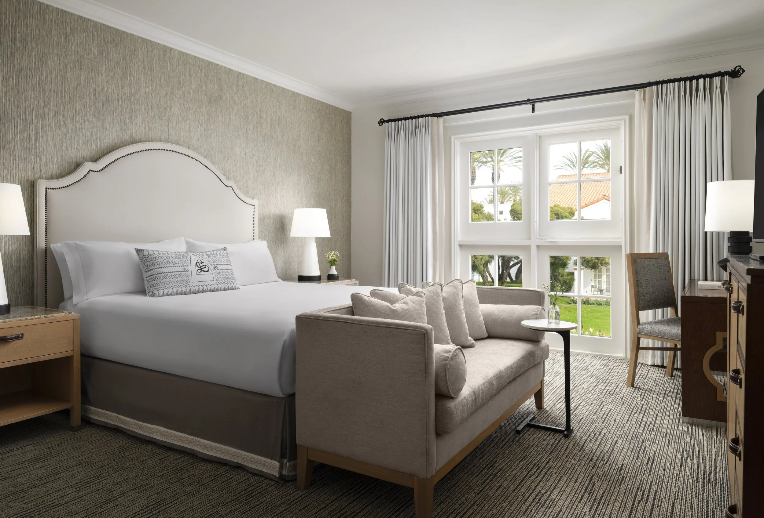 A modern hotel room with a large bed, bedside lamps, a beige sofa with pillows, a small round table with a vase, a chair, and large windows with white curtains showing palm trees outside.